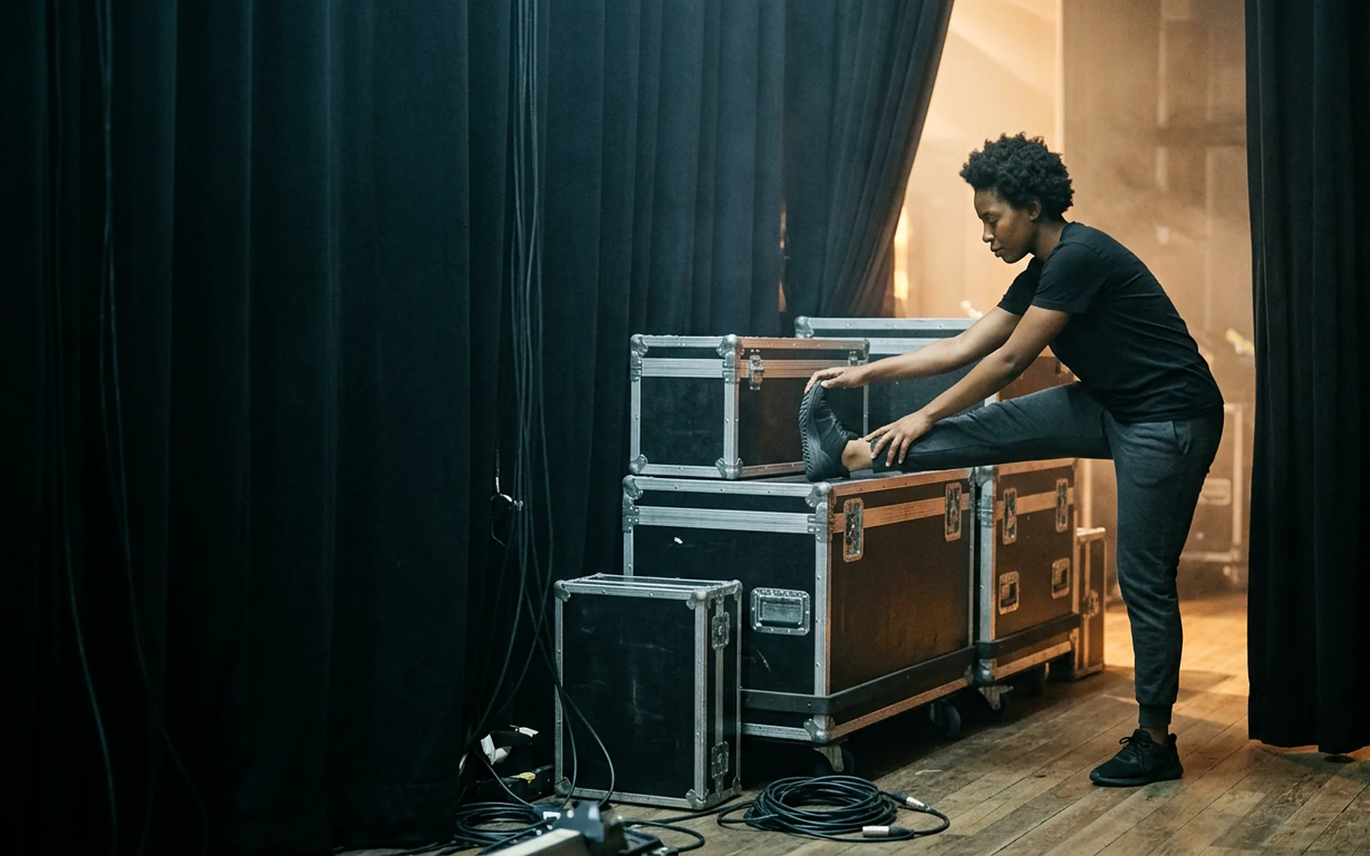 A musician stretching backstage between road cases under amber stage light